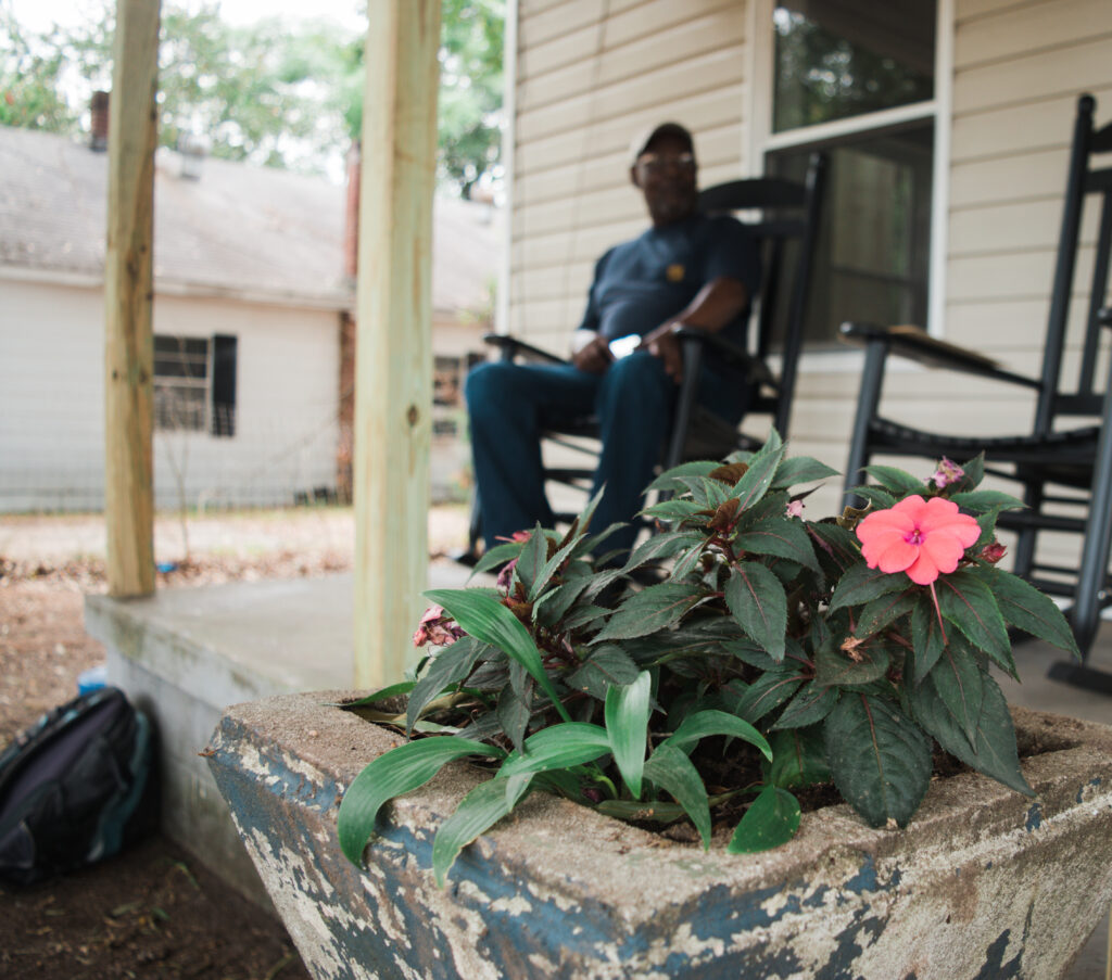 Mr. Mitchell, a 2017 homeowner, enjoys his porch while rocking on a rocking chair. Pink flowers are in the foreground.